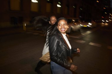 Meşgul bir şehirde, yolda koşuşturan ve gülen iki Afrikalı Amerikalı kadının flaşlı portresi. Şehir ışıkları ve hareket bulanıklığı