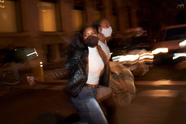 Meşgul bir şehirde gece vakti sokakta koşuşturan ve gülen yüzü maskeli iki Afro-Amerikan kadının flaşlı portresi. Şehir ışıkları ve hareket bulanıklığı