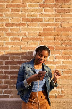 Kulaklıklı, müzik dinleyen, tuğla duvarda şarkı söyleyen ve dans eden genç Afrikalı Amerikalı kadının dikey fotoğrafı..