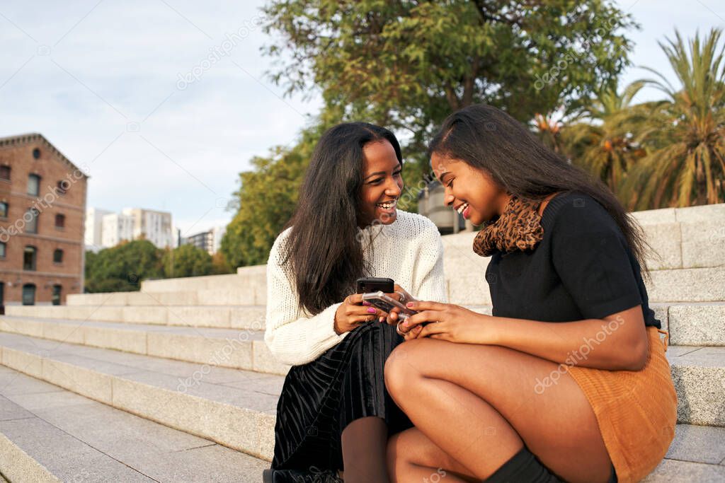 Two black girls on some stairs in a city while using a mobile phone and laughing. People and technology concept with smartphone.
