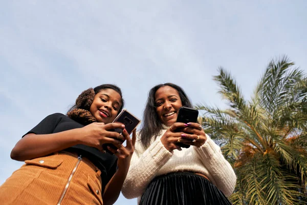 Cep telefonu kullanıp gülen iki zenci kızın alçak açılı görüntüsü. Bir tanesi kameraya bakıyor. Akıllı telefonlu insanlar ve teknoloji kavramı.