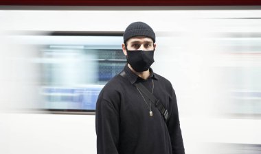 Metro peronundaki kameraya bakan maskeli bir gencin portresi. Arka planda metro kervanı hareketi.