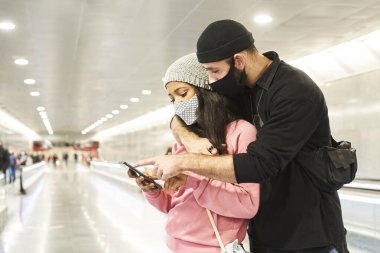 Bir metro koridorunda ya da havaalanında maskeli ve yün şapkalı genç bir çift akıllı telefondan ilginç bir şeye danışıyor..