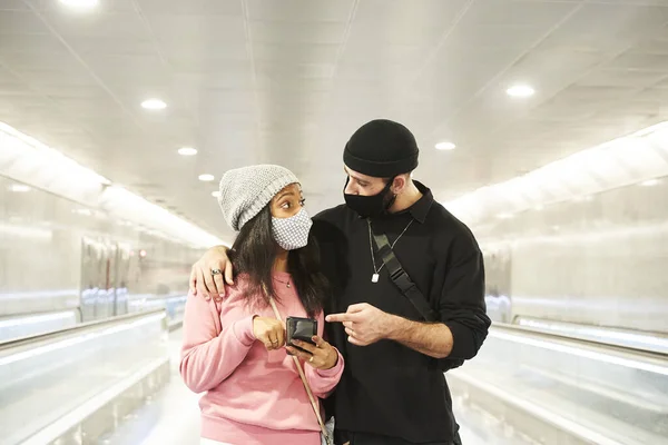 一对年轻的跨种族恋人 戴着面具和羊毛帽 走在地铁的走廊上 或者在机场用智能手机看一些有趣的东西 — 图库照片