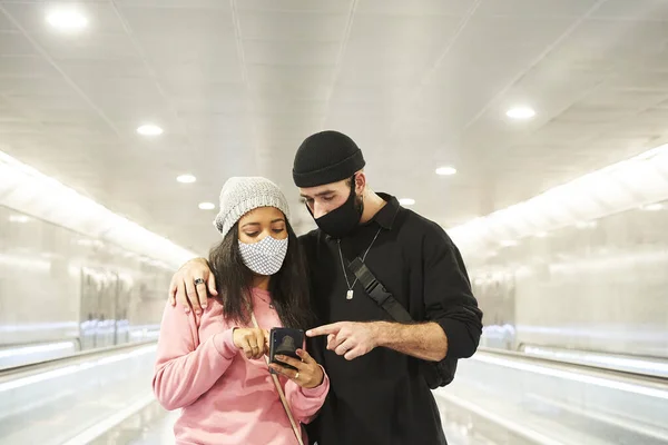 一对年轻的跨种族恋人 戴着面具和羊毛帽 走在地铁走廊上 或在机场用智能手机指着某样东西 — 图库照片