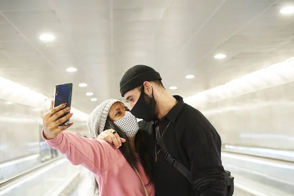 一对年轻的跨种族恋人 戴着面具和羊毛帽 在地铁或机场走廊里自拍 — 图库照片