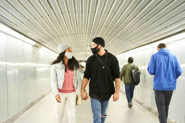 一对年轻的跨种族夫妇手牵手地走在地下通道上 年轻的黑人妇女和年轻的白人男子在公共交通上戴着面具 — 图库照片