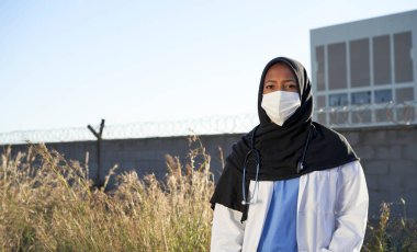 Açık havada türbanlı genç bir Müslüman doktor. Türbanlı İslami doktor dışarıda kameraya bakan fakir bir bölgede duruyor. Gönüllü doktorlar.