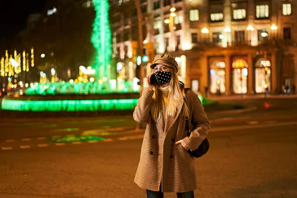 年轻的金发女郎 戴着面具 晚上在户外打电话 在背景中有许多城市的灯光 冬季气氛 — 图库照片