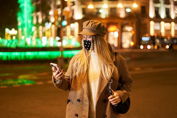 年轻的金发女郎带着面具看手机里的东西 她晚上在一座城市里 冬季气氛 — 图库照片