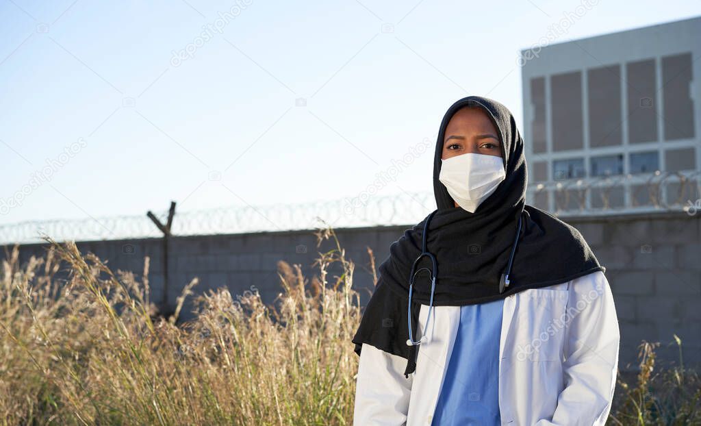 A young Muslim doctor with a headscarf outdoors. Islamic doctor with hijab is standing outside in a poor area facing the camera. Volunteer doctors.