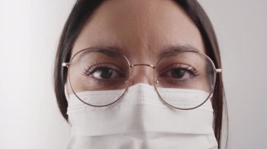 Maskeli ve gözlüklü bir kadının evinin içinde ciddi bir kameraya bakarken ki görüntüsü..