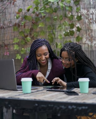 İki Afrikalı Amerikalı kadının terasta oturmuş çeşitli elektronik cihazlar üzerinde çalışırken kahve içerken dikey çekimleri. Laptop, tablet, akıllı telefon. Takım çalışması konsepti.