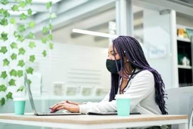 Maskeli bir iş kadını elinde laptopla elinde kahve ile ofiste oturuyor..