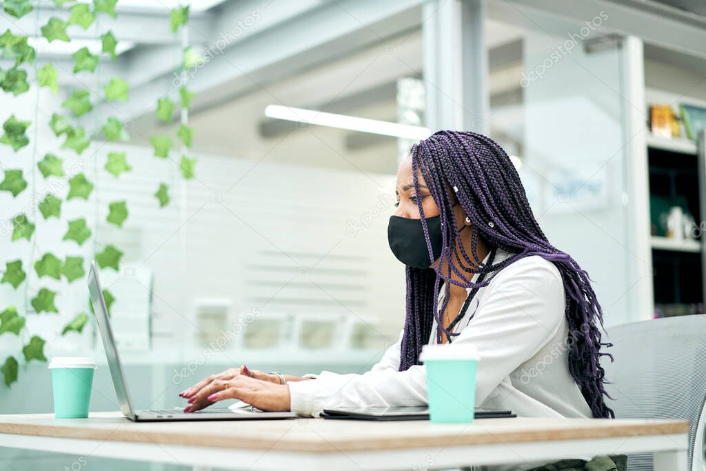 Businesswoman with a facial mask sitting at office desk holding coffee while working on laptop.