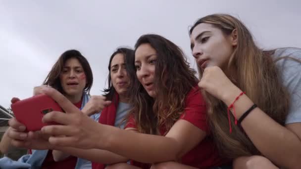 Grupo de seguidoras de un equipo de fútbol viendo un partido en streaming vestidas con camisetas rojas. Las mujeres celebran alegremente el gol con la euforia. — Vídeos de Stock