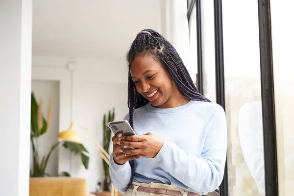 Genç Afro-Amerikalı kadın evde, pencerenin yanında akıllı bir telefon kullanıyor. Gençlik ve çevrimiçi iletişim kavramı.