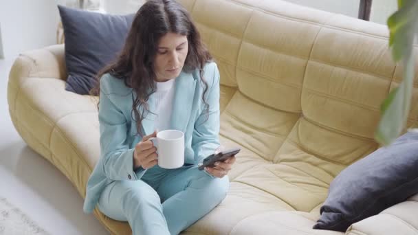 Красивая латиноамериканка во время перерыва пьет кофе, сидя на диване, пользуясь смартфоном. Работающая женщина отправляет смс с мобильного телефона, работает онлайн. 4k видео. — стоковое видео