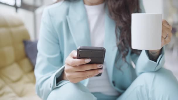 Primo piano di mani di donna d'affari irriconoscibile beve caffè seduto su un divano durante l'utilizzo di uno smartphone. La donna che lavora invia messaggi di testo dal cellulare, lavorando online. 4k video. — Video Stock