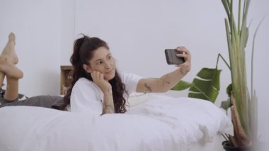 Yatakta uzanmış selfie çeken genç bir kadın. Mutluluk ve boş zaman kavramı. Etkileyici cep telefonundan çevrimiçi izleyicilerle etkileşim kuruyor.