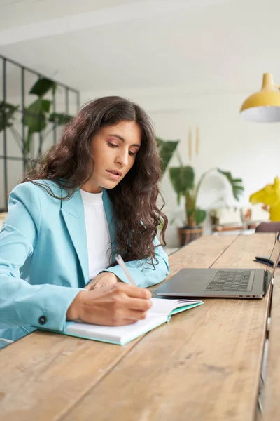Woman in office suit using laptop at home and taking notes on a notepad. Business woman working remotely from a nice modern apartment.