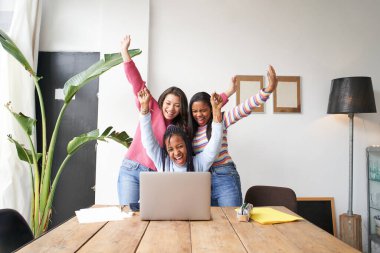 Genç kadınlardan oluşan bir çalışma ekibi iyi haberi bilgisayarın önünde kutluyor. Çok ırklı güçlü kadınlardan oluşan bir grup başarılı oldukları için birbirlerini kutluyorlar..