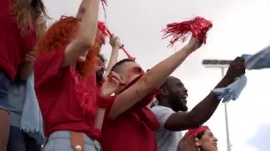 Yakınlarda farklı ırktan ve yaşlardan bir grup insan maske takarak tribünlerde futbol maçını izliyor. Erkekler ve kadınlar takım kıyafetlerini giyerler..