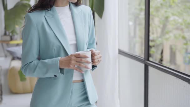 Primo piano delle mani di una giovane donna irriconoscibile in abito formale che tiene una tazza di caffè davanti alla finestra dell'ufficio. — Video Stock