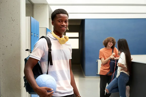 Portret Afroamerykanina patrzącego w kamerę w szkolnym korytarzu i trzymającego piłkę. — Zdjęcie stockowe