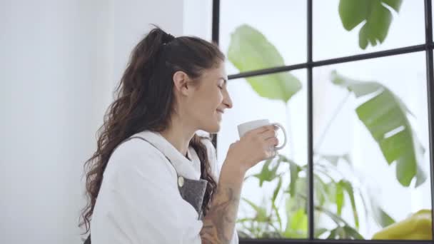 Giovane latina che guarda fuori dalla finestra godendo di una nuova sensazione giorno riposato e bere caffè a casa. Rallentatore. — Video Stock