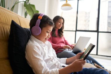 Annesi bilgisayarında uzaktan çalışırken, kulaklıklı bir çocuk evdeki kanepede tabletle oynuyor. Uzaktan çalışma ve aile uzlaşması kavramı.