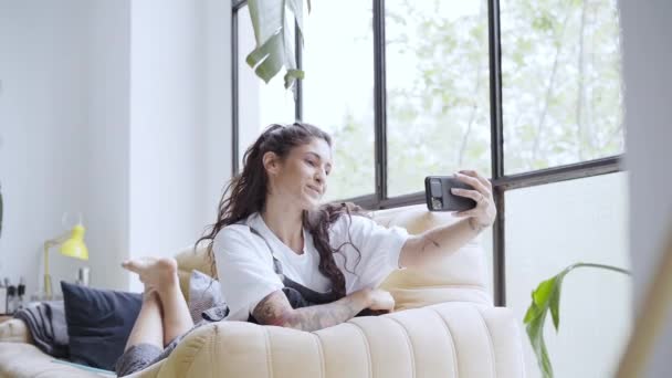 Mujer latina bastante joven toma un selfie acostado en el sofá en casa. Compartir en redes sociales y personas conectadas en línea. — Vídeos de Stock