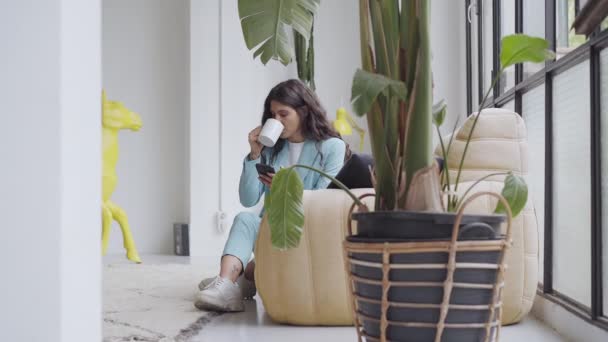 Όμορφη Λατίνα επιχειρηματίας πίνοντας καφέ σε ένα ωραίο μοντέρνο διαμέρισμα χρησιμοποιώντας ένα smartphone απολαμβάνοντας χαλαρή περιήγηση μηνύματα που σχεδιάζουν να στείλουν μηνύματα κειμένου εκ των προτέρων στο δίκτυο κινητής τηλεφωνίας. — Αρχείο Βίντεο