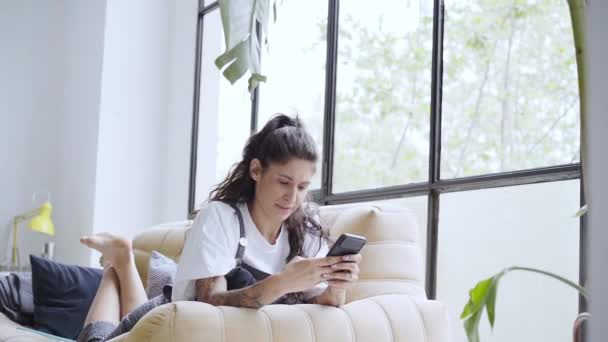 Mujer latina bastante joven usando un teléfono celular tumbado en el sofá en casa. Cara de desaprobación por algo que ve en el smartphone. Decepción por las malas noticias. — Vídeos de Stock
