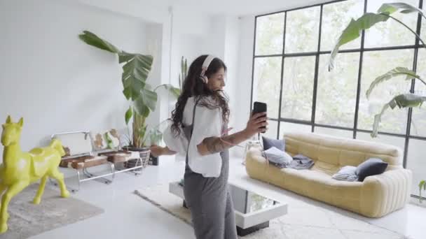 Boldog gondtalan fiatal lány táncol egyedül egy modern nappaliban egy nagy ablak hallgat zenét egy okostelefon, vicces Y-generációs lány kezében egy telefon élvezi a sikert, szórakozás otthon. — Stock videók