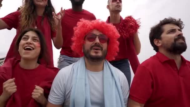 Un grupo de aficionados del equipo de fútbol celebra la victoria en las gradas con camisetas rojas mientras un fan del equipo contrario llora de tristeza. — Vídeo de stock