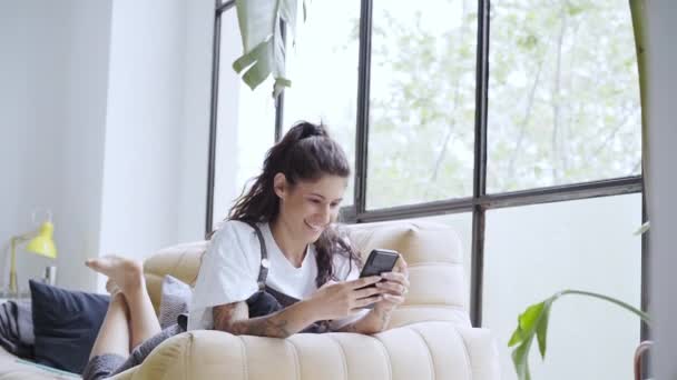 Bella giovane donna latina che utilizza un telefono cellulare sorridente sdraiato sul divano a casa. — Video Stock