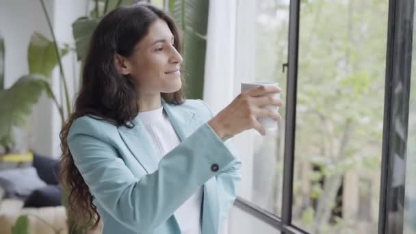 Giovane donna d'affari soddisfatta di un lavoro ben fatto rilassarsi con il suo caffè del mattino o tè, guardando fuori dalla finestra. Bella donna latina che celebra i successi durante la pausa dal lavoro in ufficio. — Video Stock