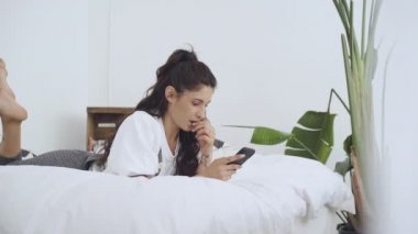 Yatakta uzanmış cep telefonu kullanan genç Latin bir kadın. Rahatlarken evlerinden birbirine bağlanan insanlar. Uygulama, mesajlaşma, online eğitim, tele-çalışma. Yavaş çekim.