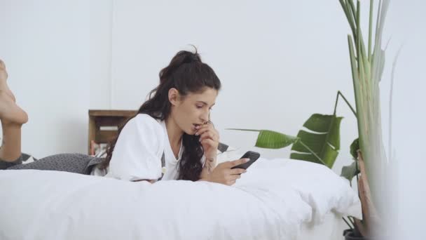 Mujer latina joven usando un teléfono móvil acostado en la cama. Personas conectadas desde casa mientras se relajan. Aplicación, mensajes de texto, educación en línea, teletrabajo. Movimiento lento. — Vídeos de Stock