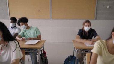 Lisedeki sınıflara koruyucu maskeler takan çok ırklı bir grup genç öğrenci katılıyor..