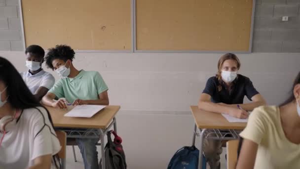 A középiskolai osztályteremben tanuló, védő maszkot viselő tinédzserek többnemzetiségű csoportja jár órákra.. — Stock videók