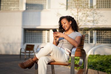 Genç, güzel bir kadın cep telefonunda oturuyor, sırıtıyor, akıllı telefon kullanıyor, gün batımında şehirde.. 
