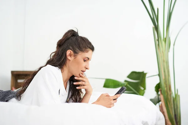 Young latina woman using a mobile phone lying on the bed. People connected from home while relaxing. App, texting, online education, teleworking.