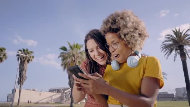 To unge kvinner ser på noe interessant på mobilen. Overrasket ansikt blant multietniske mennesker som bruker smarttelefon. – stockvideo