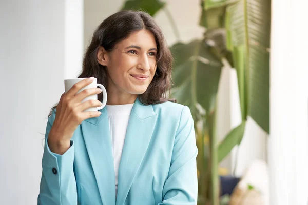 Joven mujer de negocios latina sonriente, satisfecha con un trabajo bien hecho, relajándose con su café o té de la mañana, mirando por la ventana. — Foto de Stock