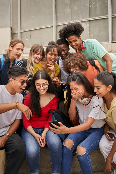 Κάθετη φωτογραφία πολυεθνικής ομάδας μαθητών που χρησιμοποιούν κινητό και γελούν. Έφηβοι που χρησιμοποιούν ένα έξυπνο τηλέφωνο, σερφάρουν στο διαδίκτυο. Διασκεδάζουμε μαζί.. — Φωτογραφία Αρχείου
