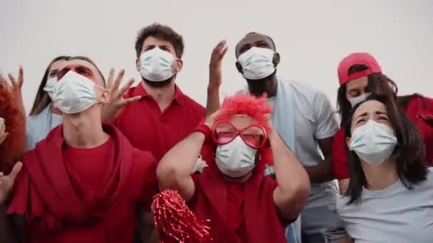 Vue de face du groupe de fans déplorant une occasion manquée pour leur équipe. A proximité, un groupe de personnes de races et d'âges différents suivent le match de football dans les gradins portant des masques. — Video