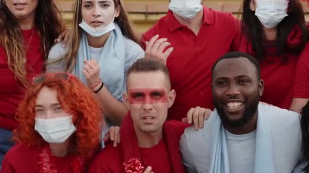 Grupo de personas de diferentes razas siguen el partido de fútbol en las gradas. Hombres y mujeres celebrando un gol vestidos con el kit de sus equipos. — Vídeos de Stock