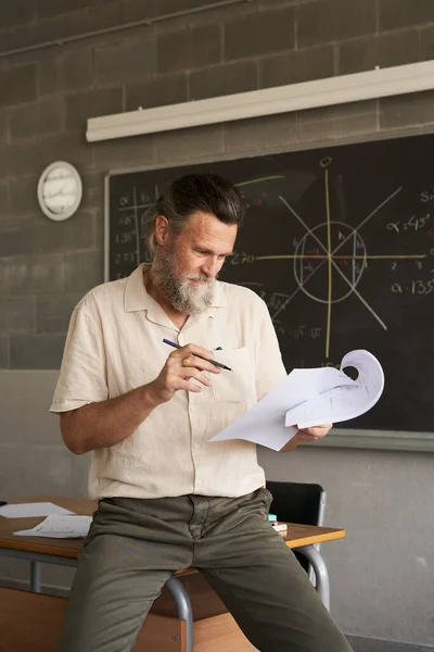 Вертикальна фотографія вчителя середнього віку з бородою виправляє іспити в класі. У руці він тримає папір і ручку . — стокове фото